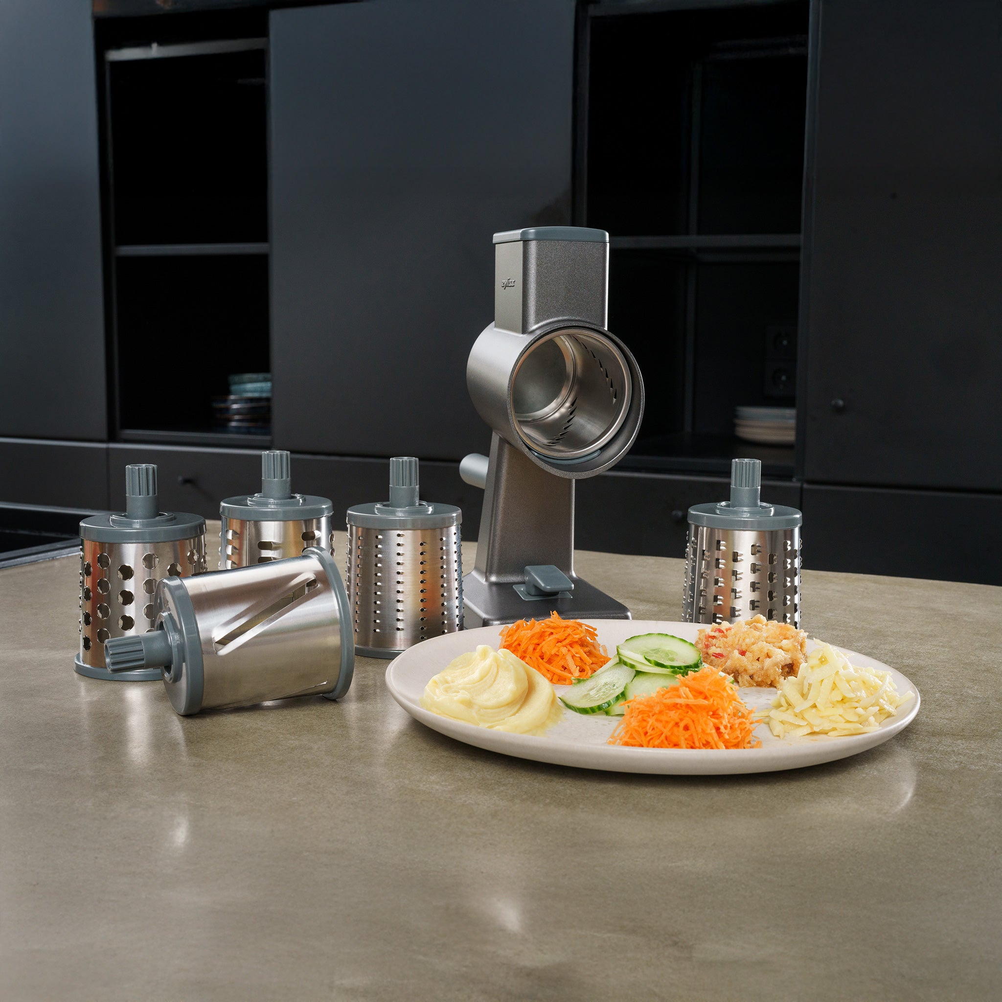 A metal rotary grater on a kitchen counter with various drum attachments. A white plate holds grated cheese, sliced cucumbers, shredded carrots, and other ingredients. Black cabinets are in the background.