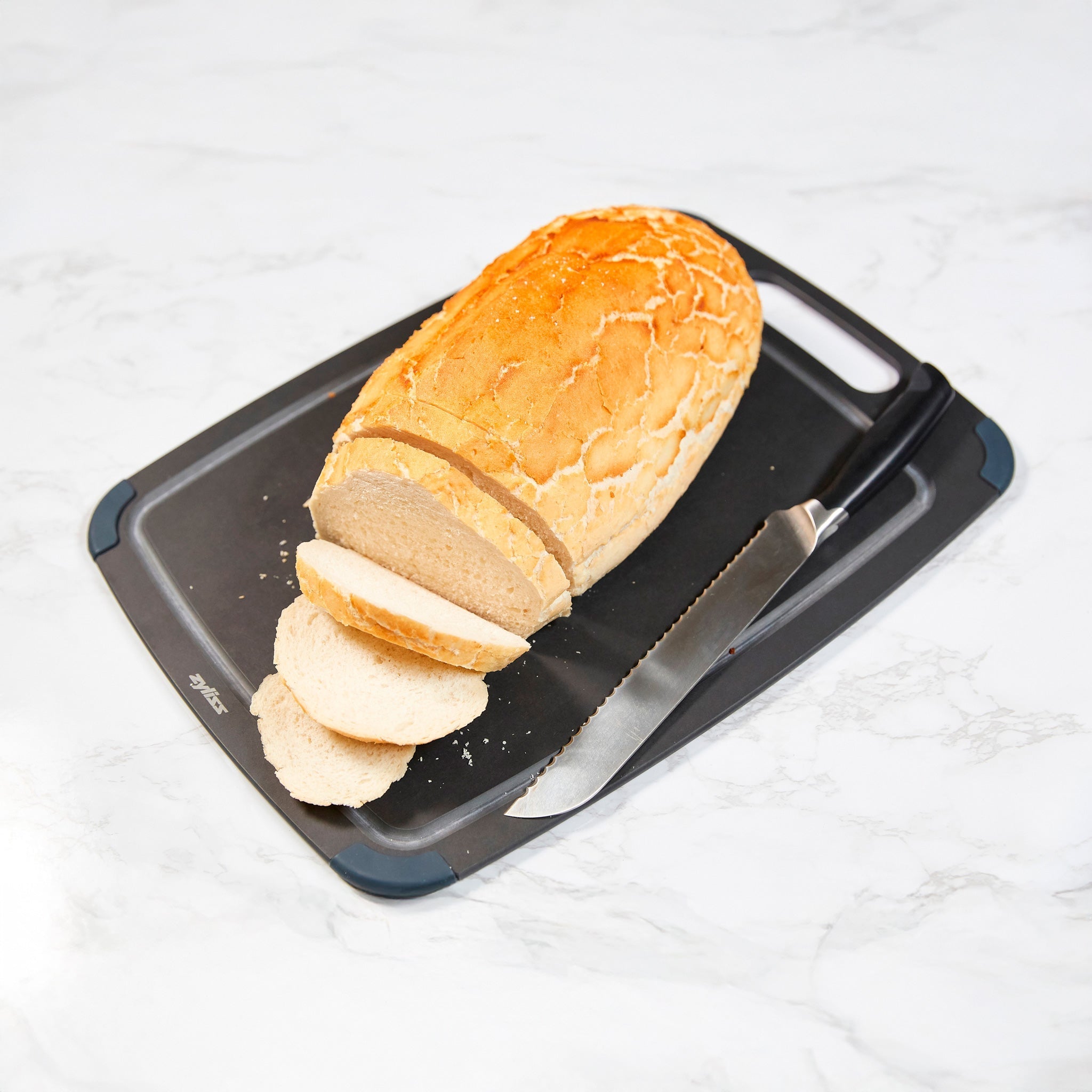 A loaf of bread with several slices cut, placed on a cutting board next to a serrated bread knife on a white marble surface.