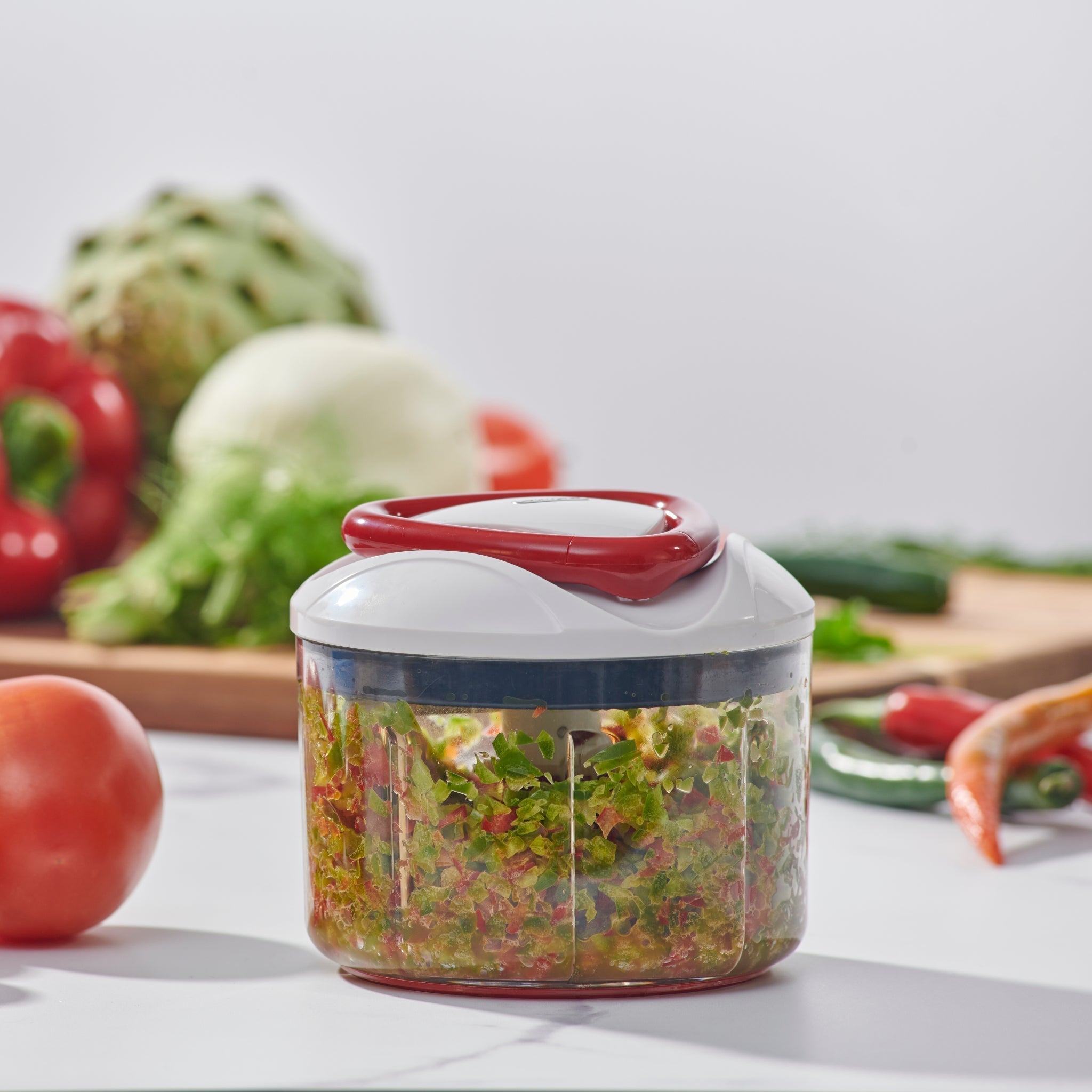 A manual food chopper filled with chopped vegetables sits on a kitchen counter, with whole vegetables and a cutting board in the background.