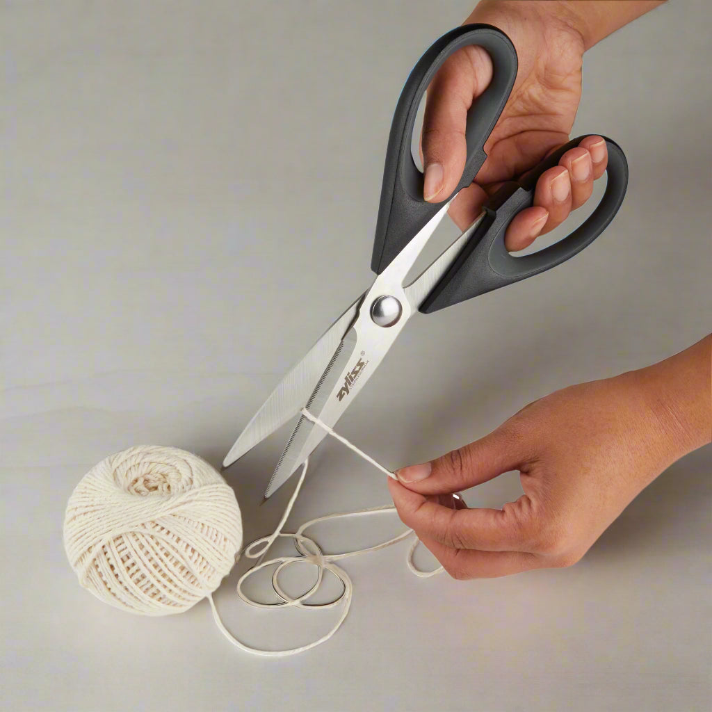 A person uses a pair of large black-handled scissors to cut a piece of white string, which is attached to a ball of twine on a light gray surface.