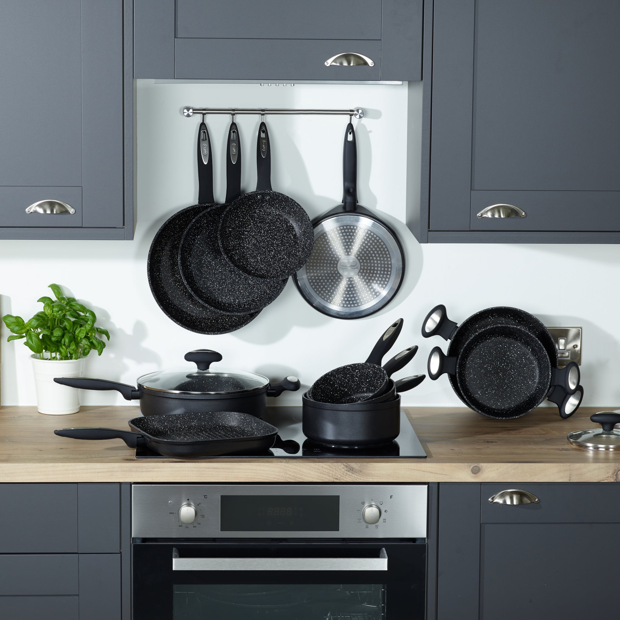 A modern kitchen with dark cabinets features black speckled cookware, including pots and pans on the stove and countertop. More pans hang on a wall rack above, next to a potted basil plant.