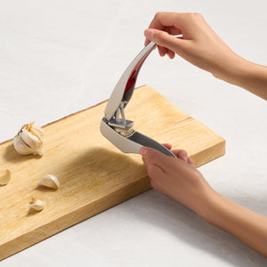A person uses the Zyliss Susi 3 Garlic Press, which is dishwasher safe and has an ergonomic handle, to crush garlic cloves on a light wooden cutting board while unpeeled garlic and extra cloves sit nearby.