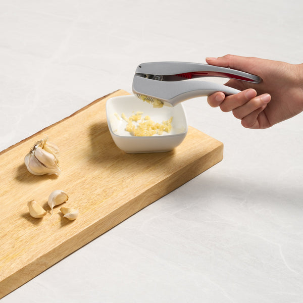 A hand uses the Zyliss Susi 3 Garlic Press with an ergonomic handle to crush garlic cloves into a small white bowl on a wooden cutting board, surrounded by whole garlic bulbs and peeled cloves.