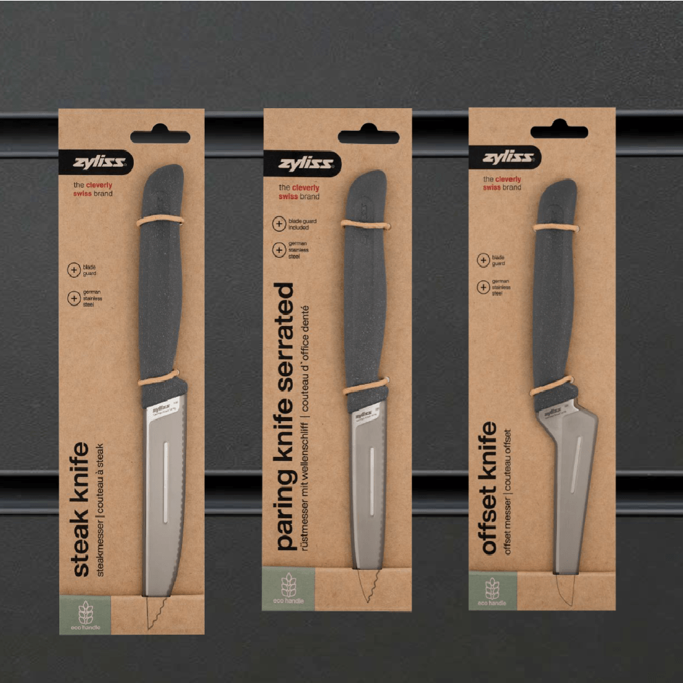Three Zyliss knives in cardboard packaging are displayed vertically: a steak knife, a serrated paring knife, and an offset knife, each with a grey handle against a dark background.