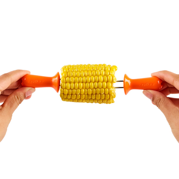 Two hands hold a piece of corn on the cob with bright orange corn holders inserted at each end against a white background.