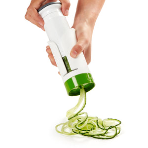A person uses a handheld spiralizer to cut a cucumber into thin, curly spiral slices, which fall onto a white surface.