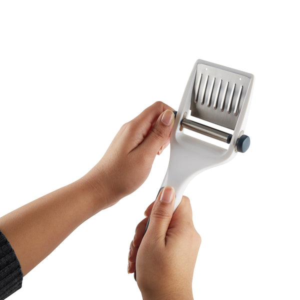 A person holds a white handheld slicer or grater with a cutting blade and metal wires, using both hands to demonstrate its use against a plain white background.