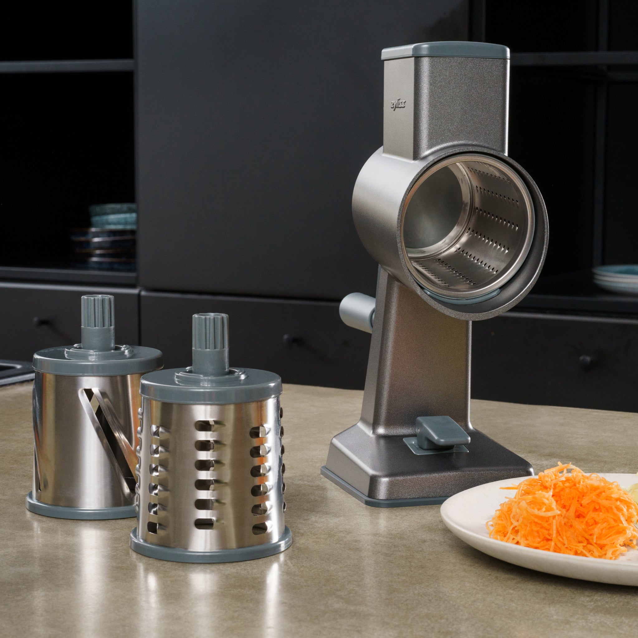 The Zyliss Gourmet Drum Grater Set rests on the counter, ready to shred fresh carrots, with stainless steel drums gleaming against sleek black cabinets. 