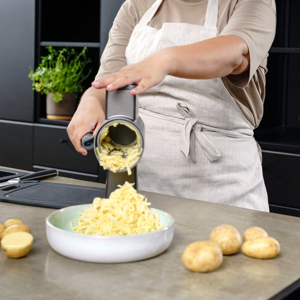 In a modern kitchen, a person uses the Zyliss Puree Drum Gourmet with its stainless steel drum to grate potatoes over a bowl. Whole and halved potatoes rest on the counter, with a potted herb adding greenery in the background. 