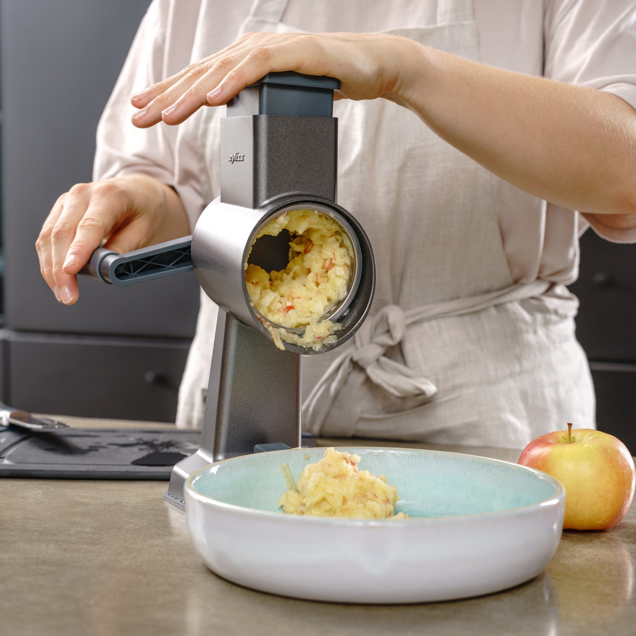Wearing an apron, a person uses the Zyliss Universal Drum Gourmet with a grating attachment to shred apples into a bowl on the kitchen counter. A whole apple beside it highlights the sleek stainless steel design of this Zyliss tool.