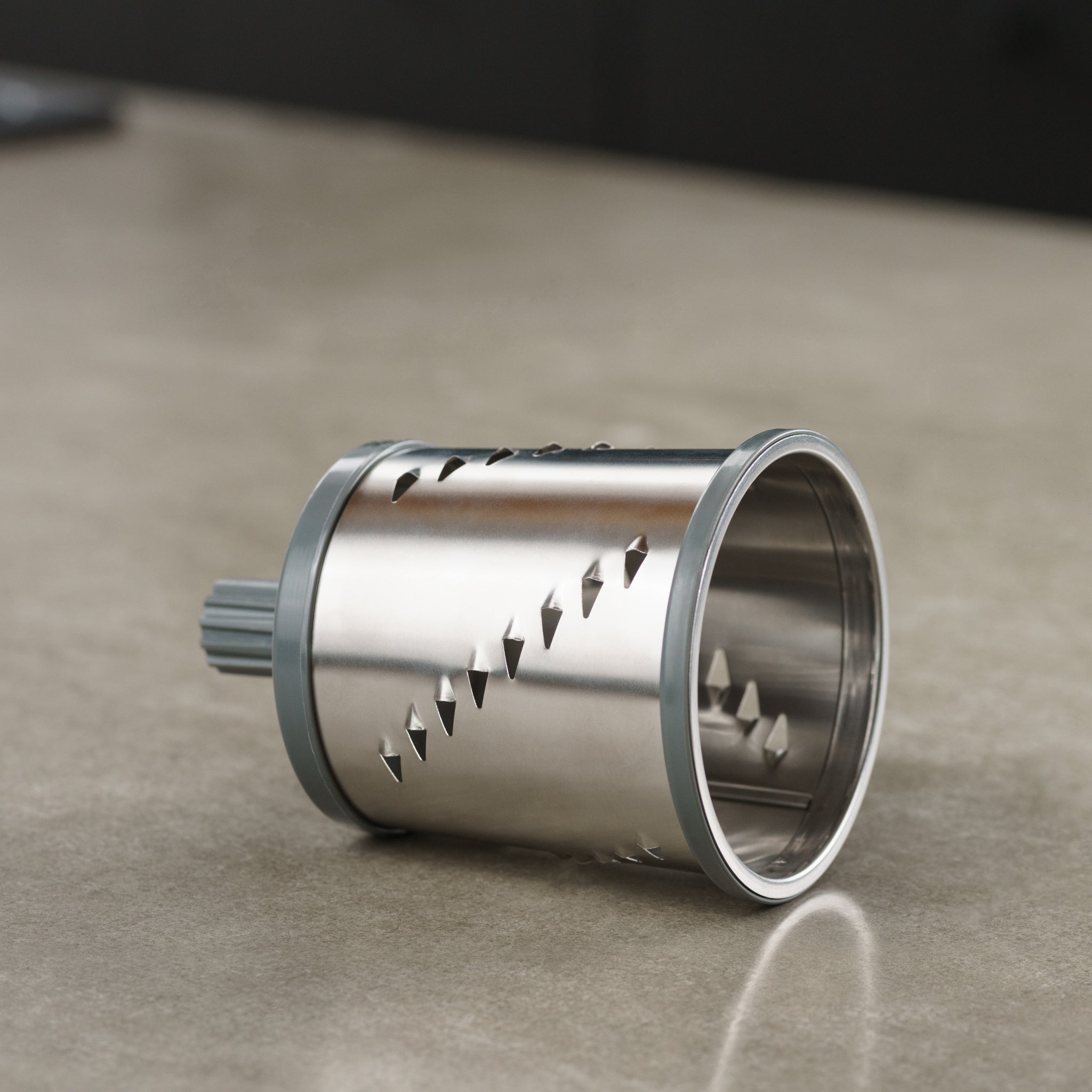 A cylindrical stainless steel rotary grater blade with sharp diagonal slots lies on a smooth, beige countertop. The blade has gray plastic edges and is designed for use in a kitchen appliance.