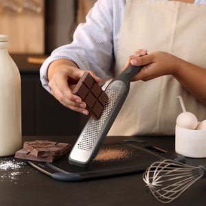 A person in an apron grates a chocolate bar over a cutting board. There are whole chocolate bars, a whisk, eggs, spilled flour, and a milk bottle on the dark kitchen counter.