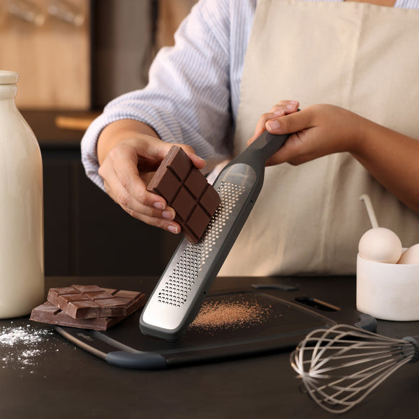 A person in an apron grates a chocolate bar over a cutting board. There are whole chocolate bars, a whisk, eggs, spilled flour, and a milk bottle on the dark kitchen counter.