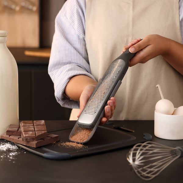 Wearing an apron, a person uses the Zyliss Rasp Grater’s ultra-sharp blade to grate chocolate onto a cutting board on the kitchen counter, surrounded by chocolate bars, eggs, a whisk, and a bottle.