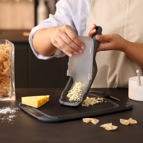 A person uses the Zyliss Coarse Grater with its ultra-sharp blade to grate cheese onto a cutting board. Nearby on the counter are a block of cheese, farfalle pasta, and a container of uncooked pasta.