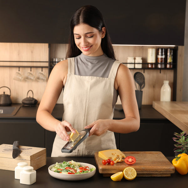 A woman in a beige apron uses the Zyliss Coarse Grater to grate cheese onto a salad in a modern kitchen. Fresh vegetables, a lemon, and a cheese block are on the cutting board as she smiles and focuses on her task.