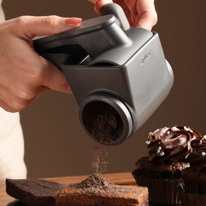 Using the Zyliss Professional Rotary Cheese Grater, a person sprinkles chocolate shavings onto a cupcake and brownies on a table.
