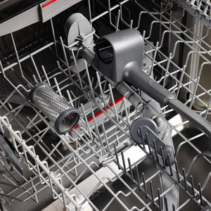 A clean dishwasher with gray racks shows only the Zyliss Professional Rotary Cheese Grater, featuring a stainless steel drum, placed horizontally on the lower rack. The spray arms and metal tines are visible.