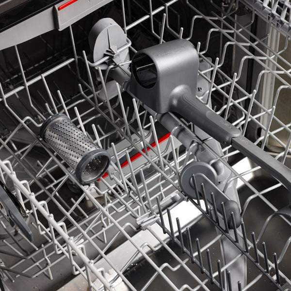 A clean dishwasher with gray racks shows only the Zyliss Professional Rotary Cheese Grater, featuring a stainless steel drum, placed horizontally on the lower rack. The spray arms and metal tines are visible.