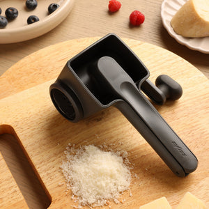 The Zyliss Professional Rotary Cheese Grater in black, featuring a stainless steel drum, sits on a wooden cutting board with grated cheese, cheese blocks, berries, and a wedge of cheese. It’s dishwasher safe for easy cleaning.