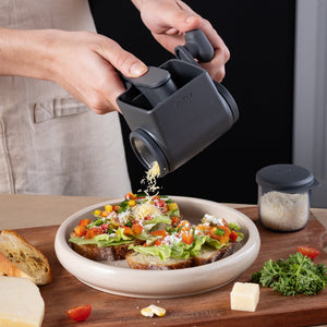 Using the Zyliss Professional Rotary Cheese Grater Set with a detachable container, cheese is grated onto open-faced vegetable sandwiches. Bread, cheese, a herb sprig, and a jar of grated cheese sit nearby on the table.