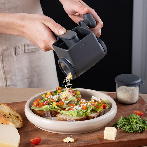Using the Zyliss Professional Rotary Cheese Grater Set with a detachable container, cheese is grated onto open-faced vegetable sandwiches. Bread, cheese, a herb sprig, and a jar of grated cheese sit nearby on the table.