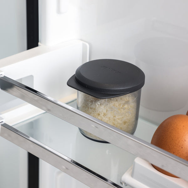 A Zyliss Professional Rotary Cheese Grater Set's glass container with a black lid, filled with grated cheese, sits on a refrigerator shelf next to a brown egg.