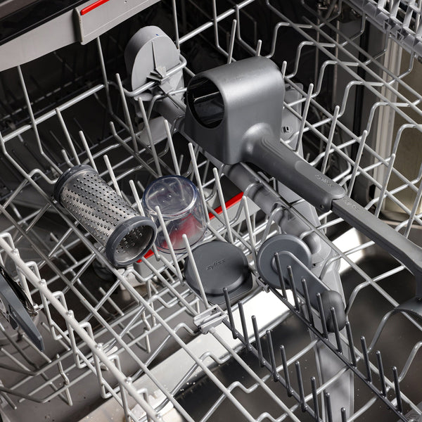 A Zyliss Professional Rotary Cheese Grater Set with detachable container and ergonomic crank, along with other gray and clear kitchen tools, sits on the top rack of an open dishwasher, ready for cleaning.