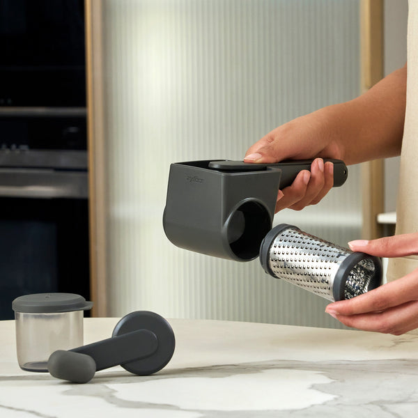 A person uses the Zyliss Professional Rotary Cheese Grater Set, holding the black ergonomic grater while removing its drum for cleaning; the detachable container and pusher are on a gray kitchen counter nearby.