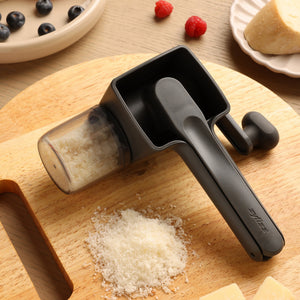 The Zyliss Professional Rotary Cheese Grater Set, in black, with a detachable container and grated cheese below, sits on a wooden cutting board near berries, cheese wedges, and a partially visible plate.