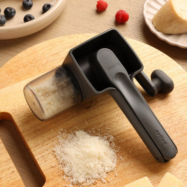 The Zyliss Professional Rotary Cheese Grater Set, in black, with a detachable container and grated cheese below, sits on a wooden cutting board near berries, cheese wedges, and a partially visible plate.
