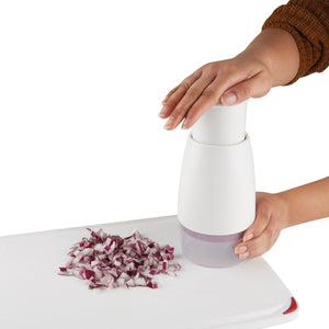 A person uses a white manual food chopper to dice red onions, which are collected in a pile on a white surface next to the chopper.
