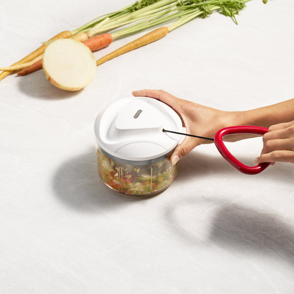 A hand pulls the red handle of the Zyliss Easy Pull Food Processor filled with chopped vegetables, while sliced carrots and a carrot top rest on a white countertop in the background.
