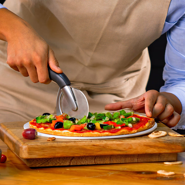 Wearing an apron, a person slices a veggie pizza topped with olives, peppers, and tomatoes on a wooden board using the Zyliss Pizza & Pastry Cutter featuring an ergonomic handle. 
