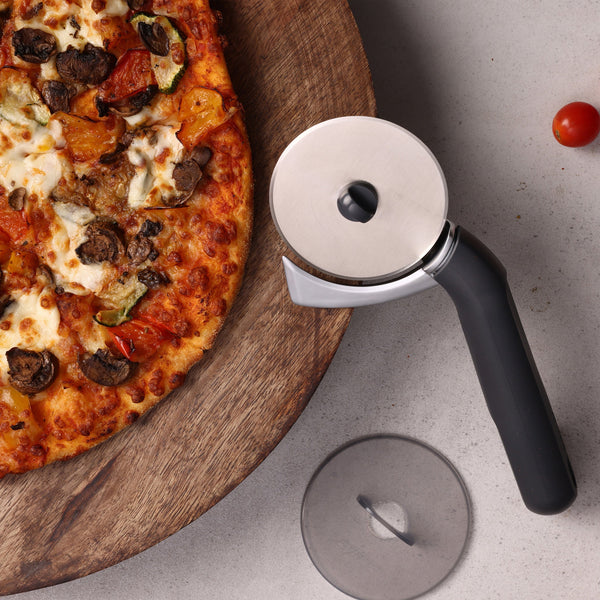 A vegetable pizza on a wooden board sits next to the Zyliss Pizza & Pastry Cutter with ergonomic handle and protective cover on a gray surface. A single cherry tomato is also visible nearby.