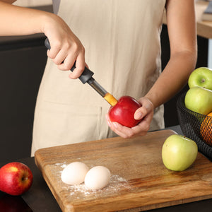 Wearing a beige apron, someone uses the Zyliss Apple Corer to remove the core from a red apple above a wooden cutting board with flour, two eggs, and apples nearby.