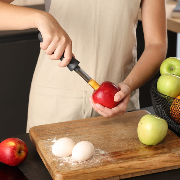 Wearing a beige apron, someone uses the Zyliss Apple Corer to remove the core from a red apple above a wooden cutting board with flour, two eggs, and apples nearby.