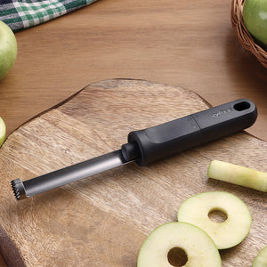 A metal apple corer with a black handle rests on a wooden cutting board, surrounded by whole green apples, a sliced apple, and apple cores.