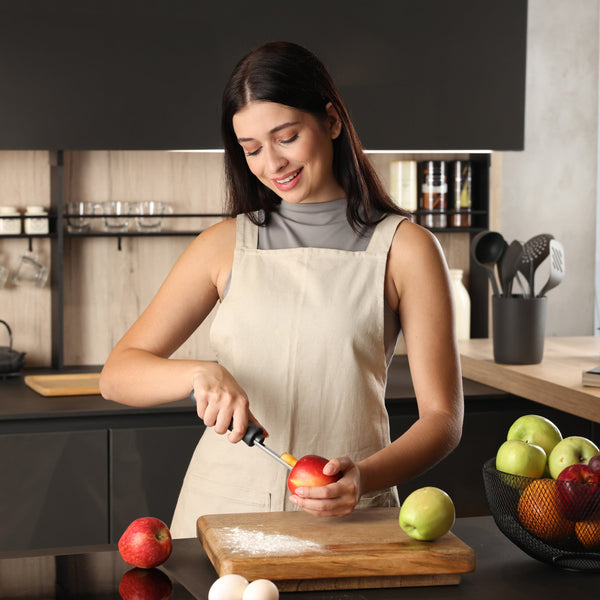 A woman in a beige apron smiles in a modern kitchen as she uses the Zyliss Apple Corer with an eco-friendly handle on a wooden cutting board. A bowl of apples, a green apple, and two eggs are also on the counter.