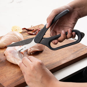 Using Zyliss All Purpose Shears with ergonomic handles and stainless steel blades, a person cuts raw chicken breast on a wooden cutting board, with lemon slice and dried chilies close by.