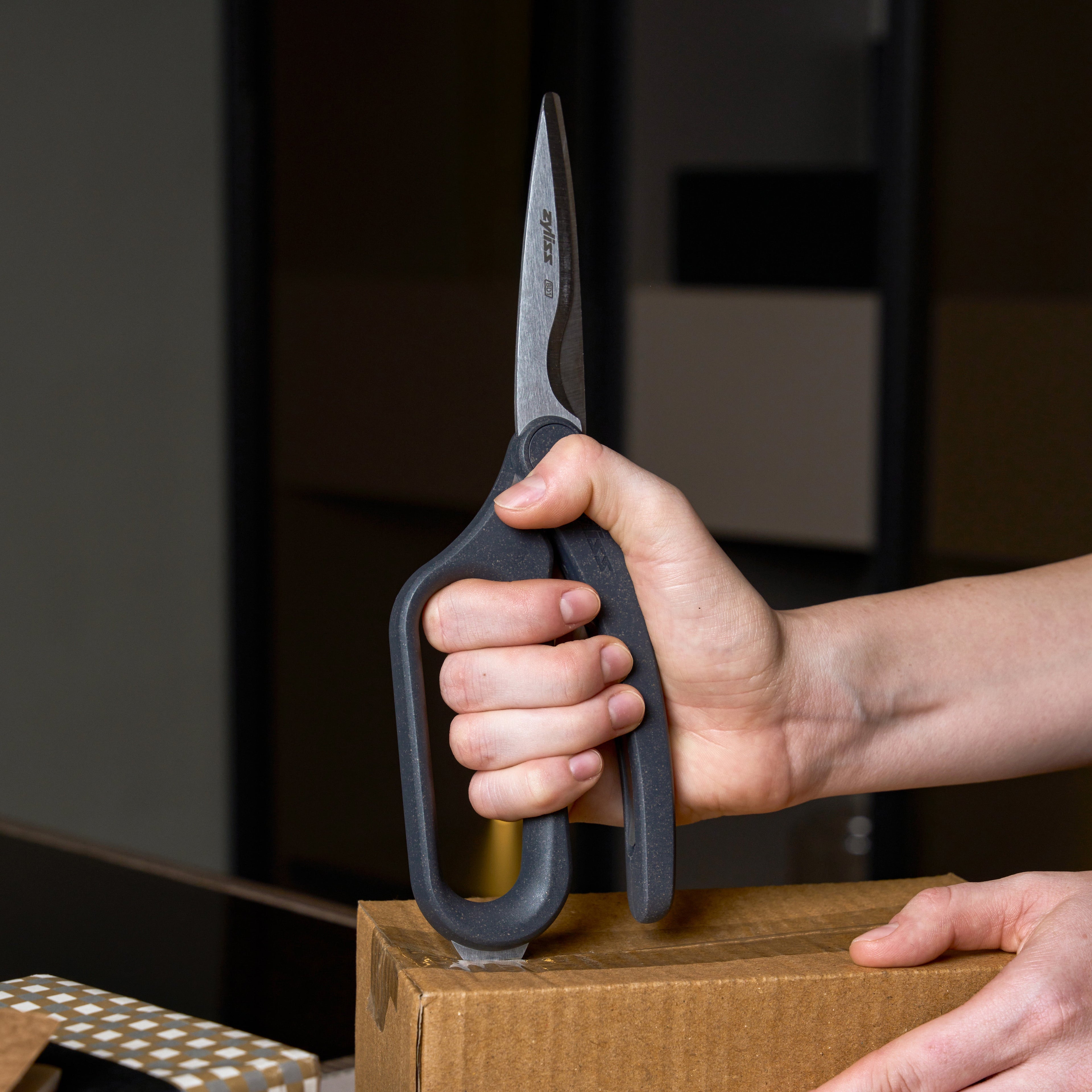 A person holds Zyliss All Purpose Shears with ergonomic handles upright, using their stainless steel blades to cut tape on a cardboard box indoors against a blurred background.