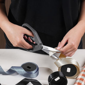 A person uses Zyliss Household Scissors with stainless steel blades to cut ivory ribbon on a table, surrounded by silver, black, and ivory rolls of ribbon.