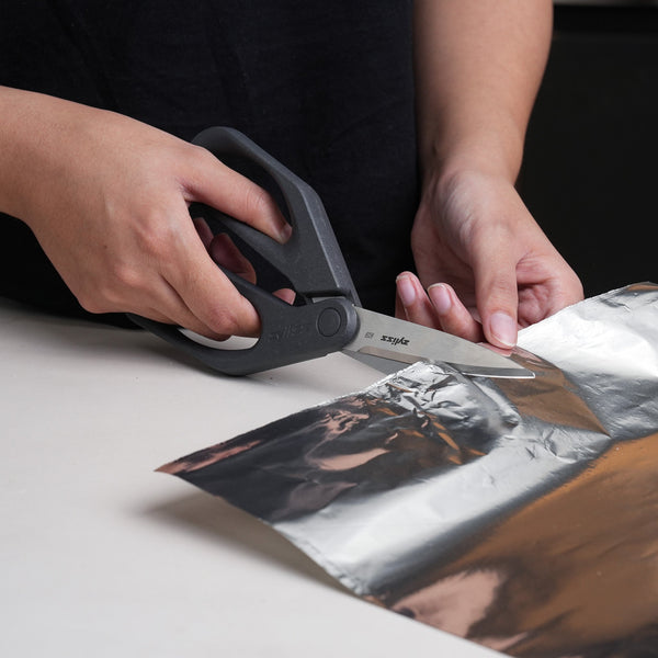 A person uses Zyliss Household Scissors with eco-friendly handles to cut aluminum foil on a white surface.
