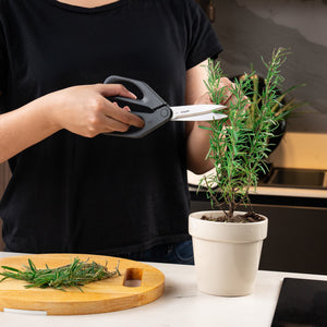 A person in a black shirt uses Zyliss Household Scissors to trim a rosemary plant in a white pot. Fresh rosemary sprigs are arranged on a wooden cutting board on a white countertop.
