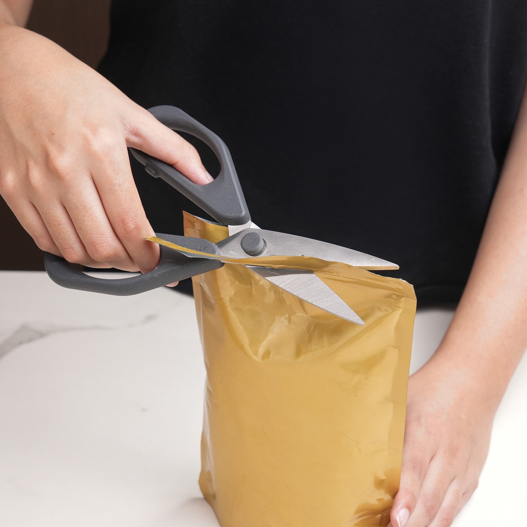 A person uses Zyliss Household Scissors Basic with stainless steel blades to cut open the top of a sealed golden-yellow plastic pouch on a white surface.