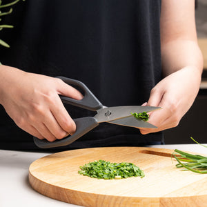 A person in a black shirt uses Zyliss Household Scissors Basic with stainless steel blades to cut fresh chives over a wooden cutting board, where chopped chives are scattered.