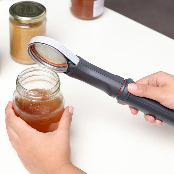Someone uses a Zyliss Jar Opener with an adjustable rubberized band to open a glass jar of honey. More jars can be seen in the background on a white surface.