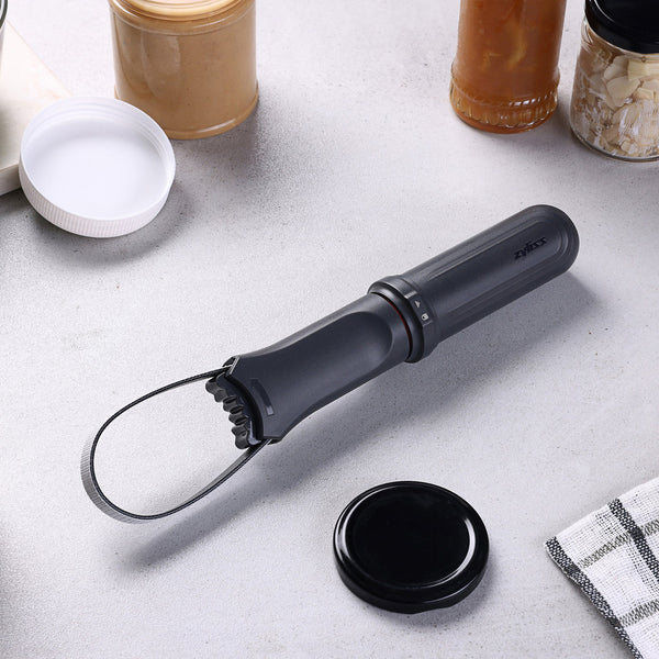 The Zyliss Jar Opener, featuring an adjustable rubberized band and handle, sits on a kitchen counter next to jars, sauces, and a checkered towel.