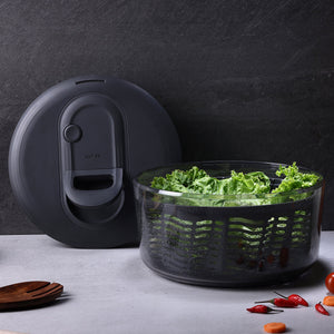 A black salad spinner with its lid resting beside it, containing fresh green leafy lettuce. The spinner sits on a countertop next to assorted vegetables and wooden utensils.