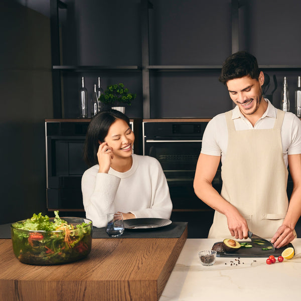 A woman smiles while sitting at a kitchen counter, watching a man in an apron slice vegetables. A large salad bowl and glass of water are on the counter. The kitchen has a modern, dark interior.